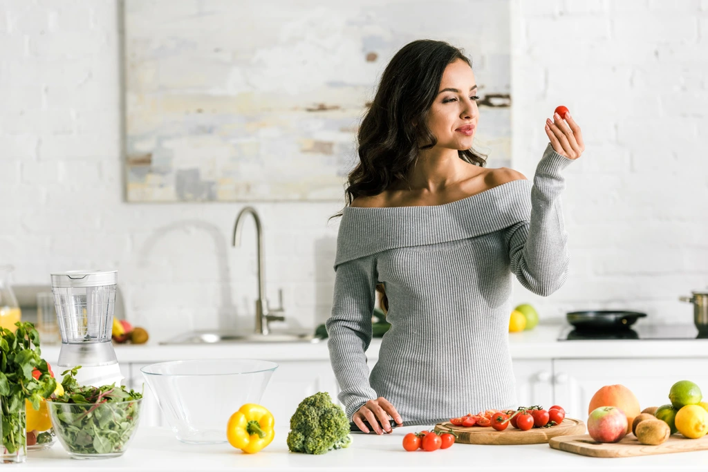 Elena Valdez, founder of One Dish Universe, smiling as she selects a fresh cherry tomato in her bright, modern kitchen.