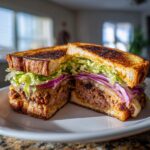 A thick, grilled Leftover meatloaf sandwich cut in half, showing layers of meatloaf, melted cheese, red onion, and lettuce.