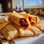 A pile of freshly made tamales wrapped in corn husks, one is partially opened revealing a rich, dark red filling.