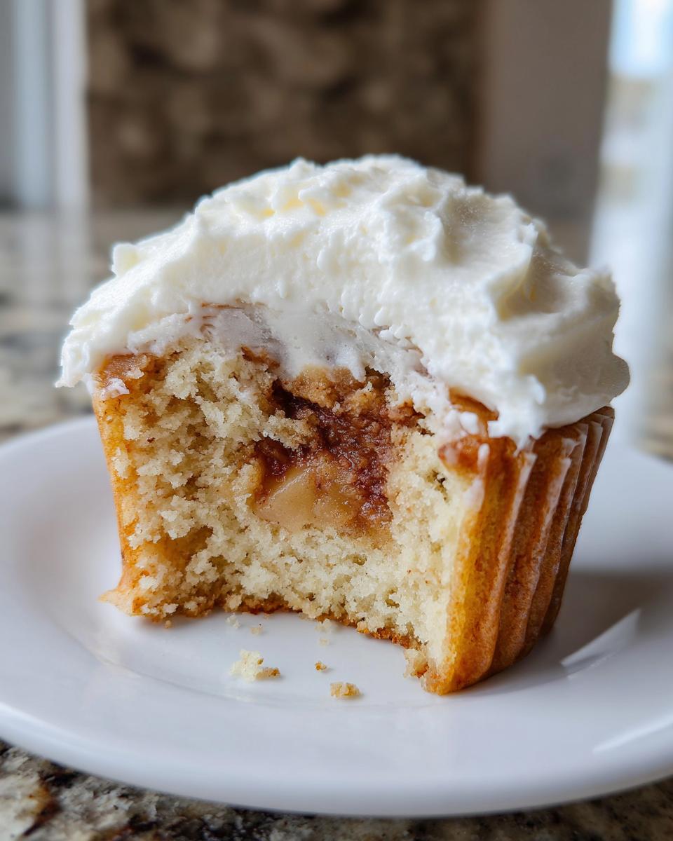 A cross-section view of an Apple pie cupcakes showing the cake texture and apple filling, topped with white frosting.