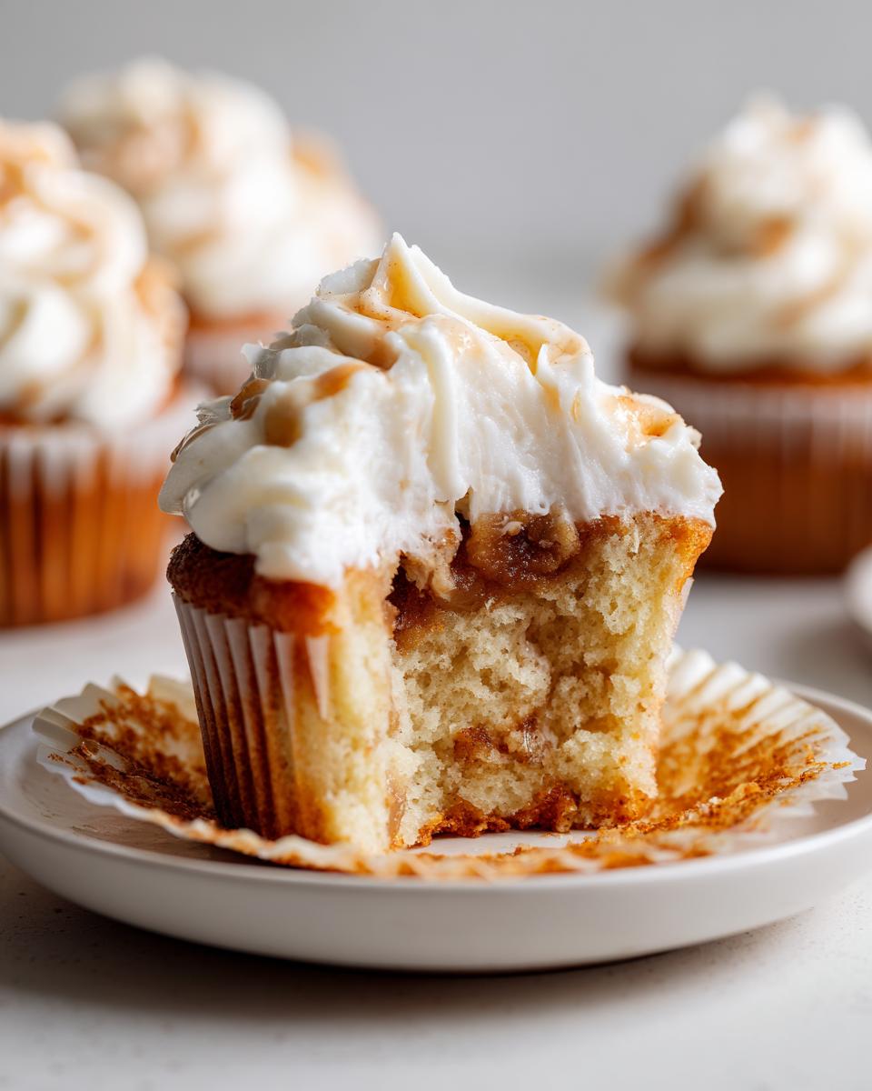 Close-up of an Apple pie cupcakes with a bite taken out, showing the filling, white frosting, and caramel drizzle.