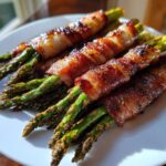 Close-up of several bundles of crispy, caramelized bacon wrapped asparagus spears resting on a white plate.