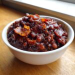 Close-up of a white bowl filled with rich, dark BBQ baked beans topped with crispy bacon pieces.
