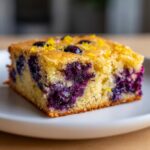 A close-up of a single, moist square of blueberry lemon blondies, studded with deep purple blueberries and topped with bright lemon zest.