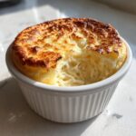 A golden-brown, perfectly risen cheese soufflé with a light, airy interior visible, served in a white ramekin.