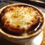 A rich, bubbling bowl of French onion soup topped with a thick layer of melted, browned cheese over crouton.