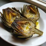 Three halves of Classic grilled artichokes, charred and drizzled with olive oil and black pepper on a white plate.