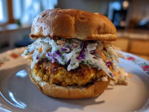 A close-up of a golden Coconut shrimp burger patty topped generously with colorful coleslaw on a toasted bun.