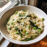 A close-up of a creamy bowl of Asparagus Risotto topped with shaved Parmesan cheese, peas, and black pepper.