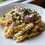 A close-up of creamy Esquites pasta salad featuring fusilli pasta, charred corn, crumbled white cheese, and red onion slices.