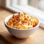 A white bowl filled with creamy, orange Pimento cheese, featuring visible red pimento pieces, set on a wooden surface.