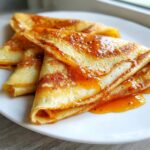 Close-up of folded Crepe Suzette drizzled generously with a glistening, amber-colored orange sauce on a white plate.