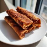 A stack of golden brown, cinnamon-sugar coated Apple pie taquitos resting on a white plate.