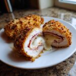 Close-up of a cut Chicken Cordon Bleu recipe piece showing melted cheese oozing out, wrapped in ham, with a crispy breaded exterior.