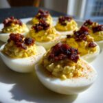 A plate of creamy deviled eggs with candied bacon topping, sprinkled with paprika.
