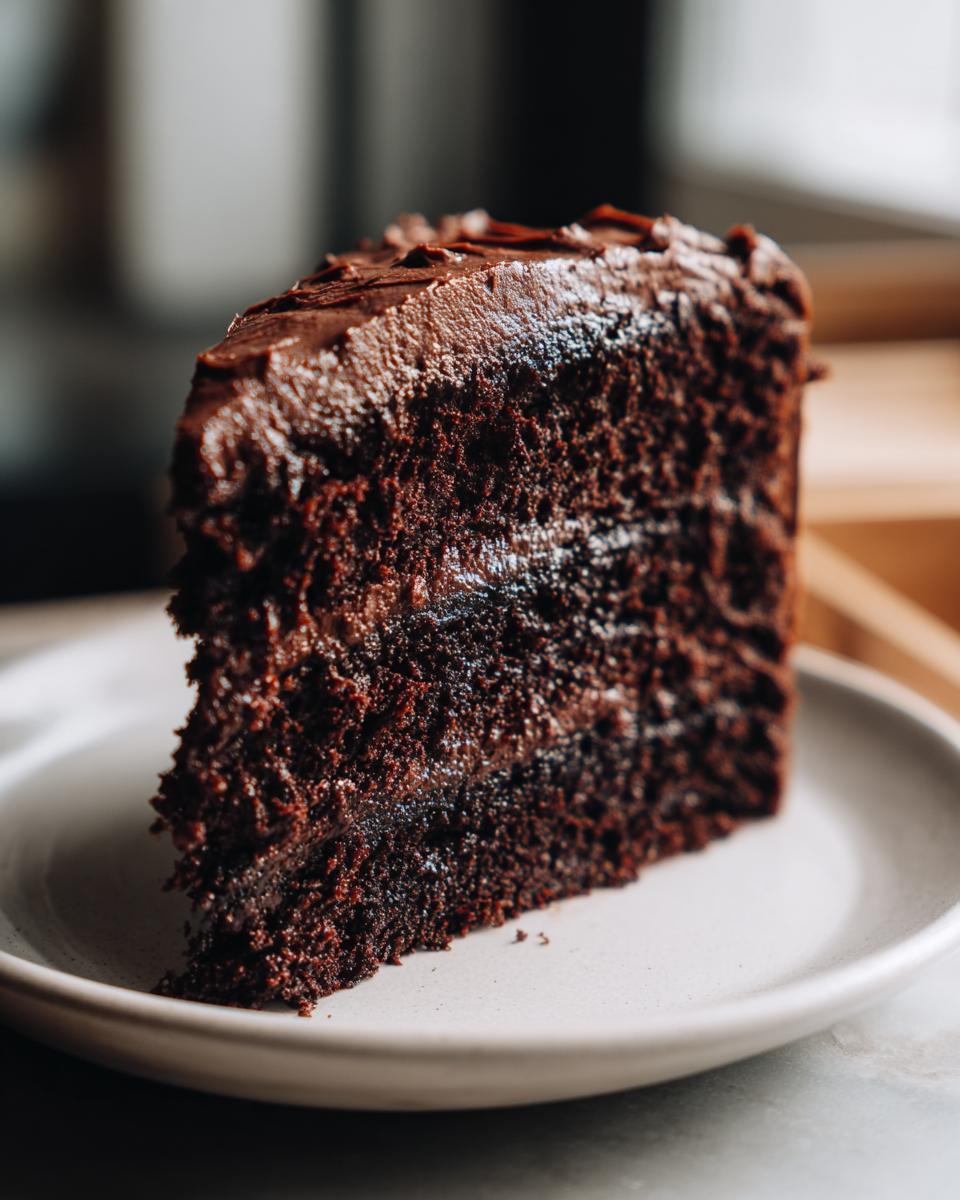 A moist, dark slice of Devil's food cake layered with thick chocolate frosting on a light plate.