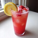 A tall glass filled with bubbly pink strawberry lemonade, ice, sliced strawberries, and a lemon wheel garnish.