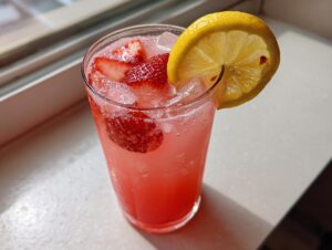 Close-up of a tall glass filled with pink strawberry lemonade, ice, fresh strawberry slices, and a lemon wheel garnish.