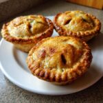 Close-up of three freshly baked, golden brown mini apple pies with crimped edges served on a white plate.