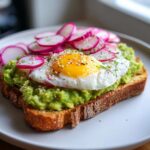 Close-up of gourmet avocado toast topped with mashed avocado, a sunny-side-up egg, sliced radishes, and sesame seeds.