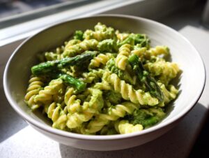Close-up of fusilli pasta coated in creamy green sauce, featuring bright green asparagus spears, served in a light bowl.