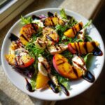 A close-up of a vibrant Grilled peach burrata salad featuring grilled peach slices, creamy burrata, and mixed greens drizzled with balsamic glaze.