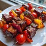 Close-up of perfectly charred and glazed grilled steak kabobs featuring beef cubes, cherry tomatoes, bell peppers, and onions on a white plate.