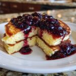 A stack of fluffy Lemon Ricotta Pancakes cut open to show the texture, topped generously with dark berry compote.