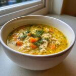 A close-up of a white bowl filled with bright yellow Mediterranean lemon chicken soup, featuring shredded chicken, carrots, orzo pasta, and fresh dill.