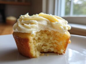 A close-up of a lemon cupcake with a bite taken out, showing the moist crumb and thick layer of cream cheese frosting.