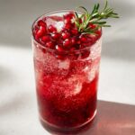 Close-up of a tall glass filled with a vibrant red Pomegranate spritz mocktail, ice, and topped with fresh pomegranate seeds and a sprig of rosemary.