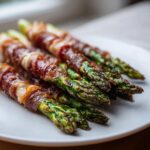 Close-up of perfectly cooked Prosciutto wrapped asparagus bundles resting on a white serving plate.