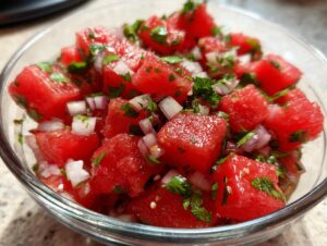 Close-up of vibrant red, cubed watermelon salsa mixed with diced red onion and fresh green herbs in a clear glass bowl.