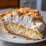 A thick slice of creamy Coconut Cream Pie with a graham cracker crust, topped with whipped cream and toasted coconut flakes.