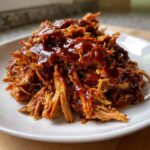 A close-up of tender, shredded Slow Cooker Pulled Pork generously coated in rich, glossy barbecue sauce on a white plate.