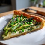 A close-up of a slice of Spring vegetable tart featuring bright green asparagus spears and peas baked into a creamy filling.