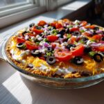 A vibrant Star spangled layer dip topped with tomatoes, olives, cheese, and onions in a clear glass pie dish.