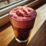 Close-up of a glass topped with thick, vibrant pink strawberry cold foam catching bright sunlight.