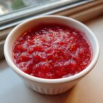 A small white ramekin filled with vibrant, chunky strawberry rhubarb sauce, sitting near a window.