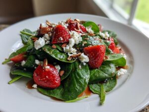 A close-up of a fresh Strawberry spinach salad topped with crumbled feta cheese, sliced almonds, and balsamic glaze.