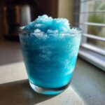 A vibrant, icy blue milk slush piled high in a clear glass, catching sunlight on a countertop.