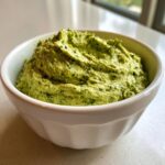 A close-up of vibrant green Greek goddess dip, textured and speckled with herbs, served in a white bowl.