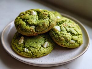 A stack of three vibrant green White Chocolate Chip Matcha Cookies resting on a speckled, light-colored ceramic plate.